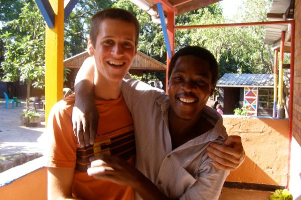 Ein Zivildienstpflichtiger während eines Freiwilligendienstes und ein junger Mann stehen nebeneinander unter einer überdachten Holzterrasse, jeder hat einen Arm um die Schulter des Anderen gelegt
