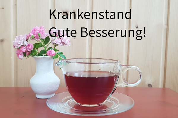 Symbolfoto mit einer Tasse aus Glas mit Tee, links daneben steht eine weiße Vase mit rosaroten Blumen. Die Tasse Tee steht auf einem roten Tisch.