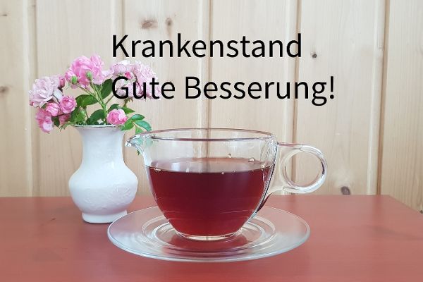 Symbolfoto mit einer Tasse aus Glas mit Tee, links daneben steht eine weiße Vase mit rosaroten Blumen. Die Tasse Tee steht auf einem roten Tisch.