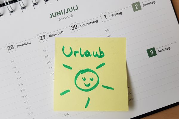Kalenderblatt mit Überschrift Juni/Juli, auf dem Kalenderblatt klebt ein gelbes Post-It mit der Aufschrift "Urlaub" und einer aufgemalten lachenden Sonne
