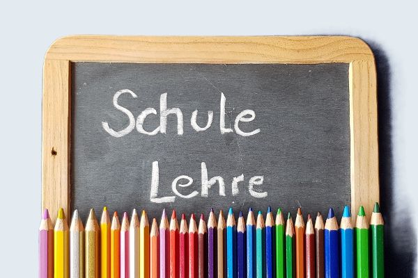 Auf einer kleinen Schultafel stehen die Begriffe Schule und Lehre. Unterhalb der Schultafel liegen nebeneinander Buntstifte.