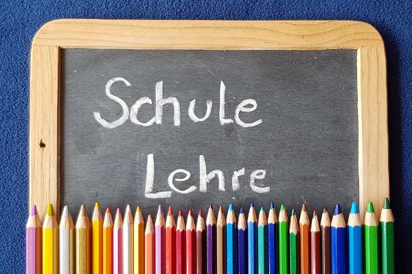 Auf einer kleinen Schultafel stehen die Begriffe Schule und Lehre. Unterhalb der Schultafel liegen nebeneinander Buntstifte.