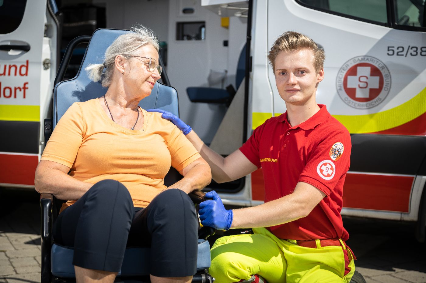 Das Foto zeigt einen Zivildiener, eine Dame, die in einem Sessel sitzt und im Hintergrund ein Rettungsauto.
