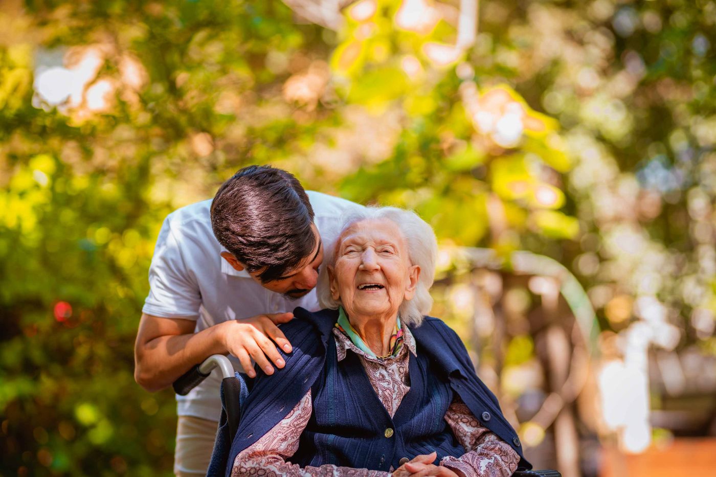 Eine Seniorin sitzt in einem Rollstuhl. Sie lacht. Ein Zivildiener steht hinter dem Rollstuhl, er hat seinen Kopf zur Dame geneigt und hört ihr zu.