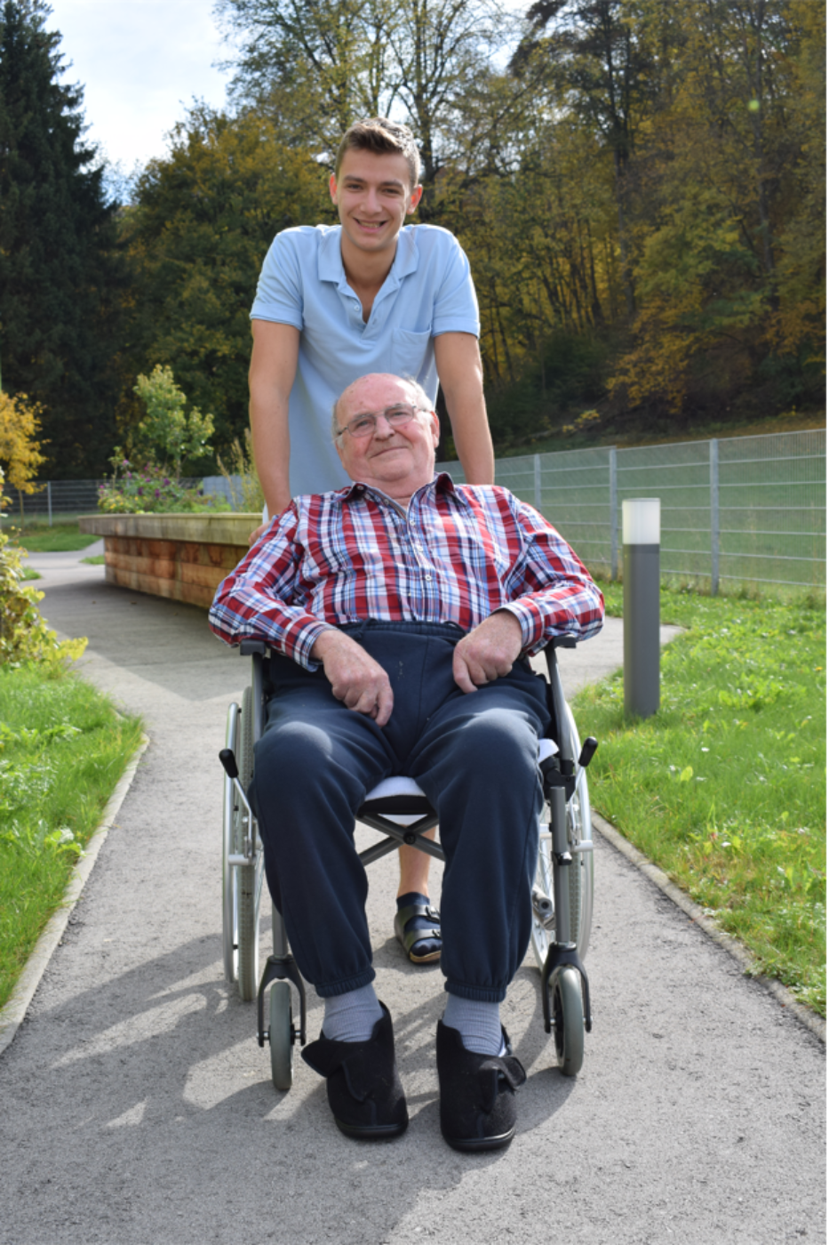 Ein Zivildiener schiebt einen Rollstuhl, in dem ein älterer Mann sitzt, auf einem Weg im Park