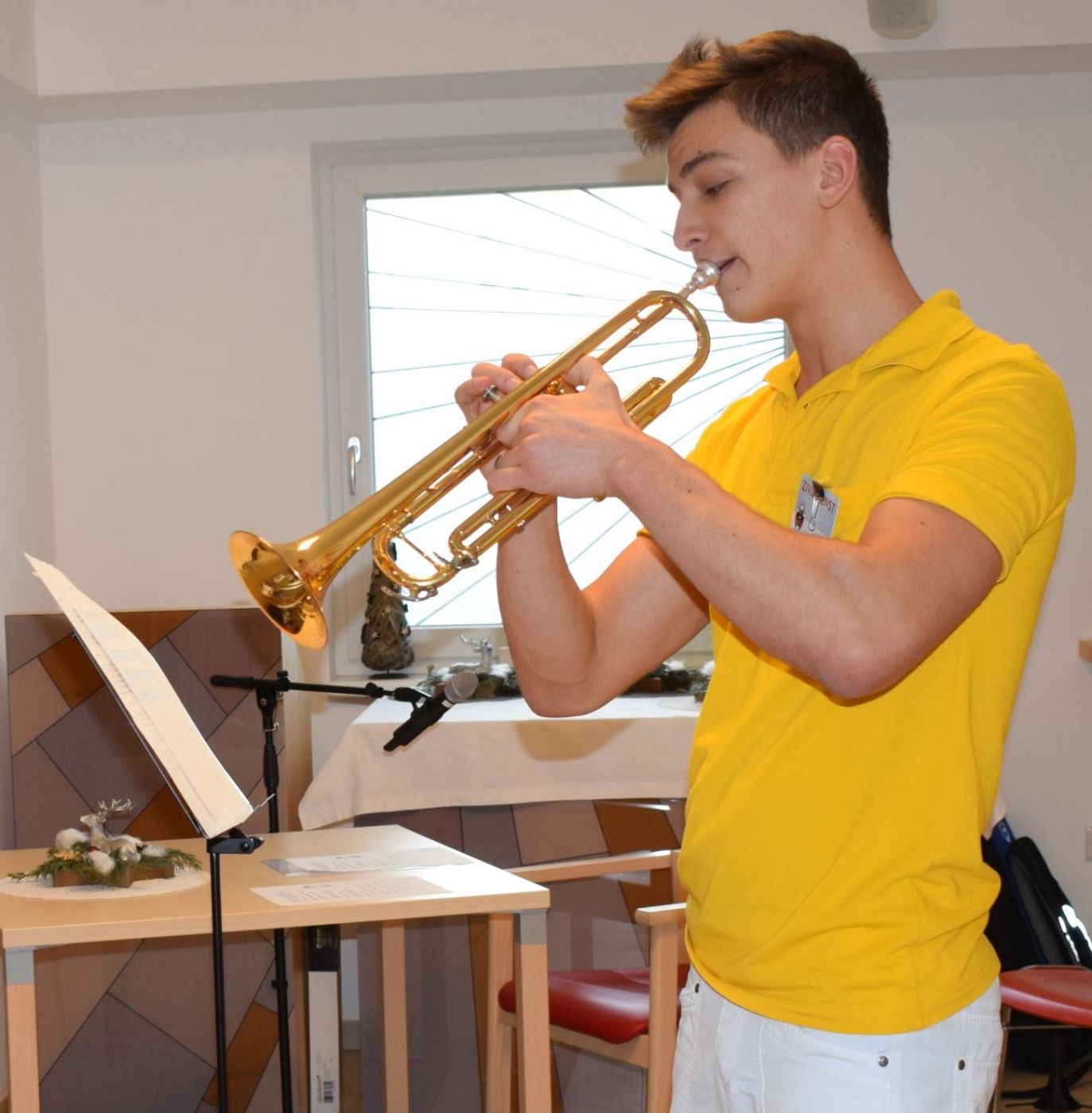 Ein Zivildiener spielt im Seniorenheim mit einer Trompete, er trägt ein gelbes T-Shirt und eine weiße Hose.