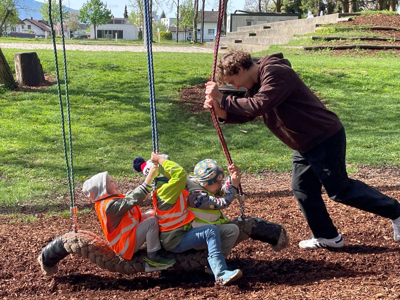 Drei Kinder sitzen auf einer Schaukel im Freien, ein Zivildiener schiebt leicht an den Seilen der Schaukel, um diese zu bewegen.