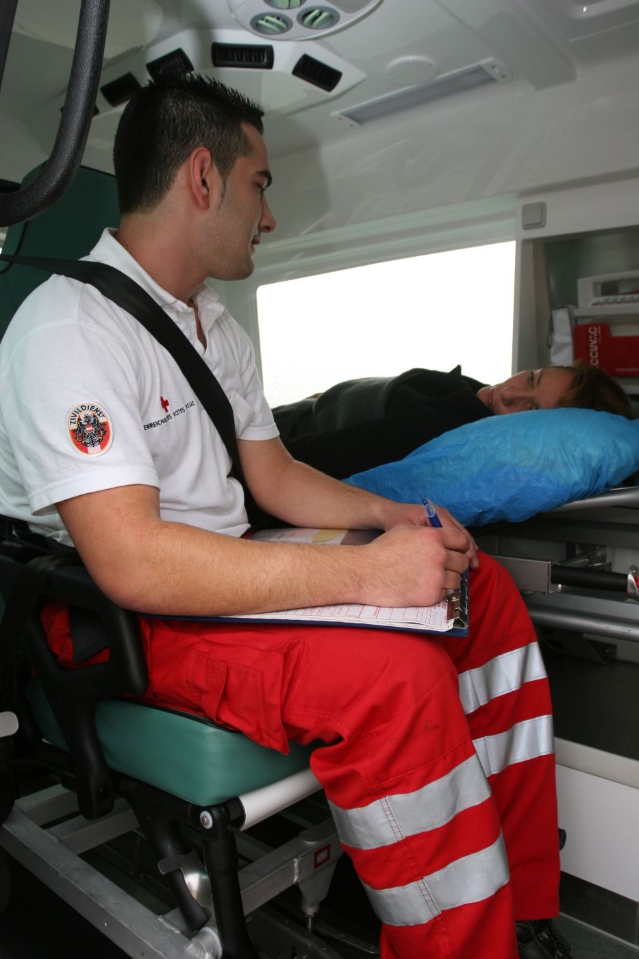 Ein Zivildiener sitzt in einem Rettungswagen, eine Patientin liegt neben dem Zivildiener im Rettungswagen.