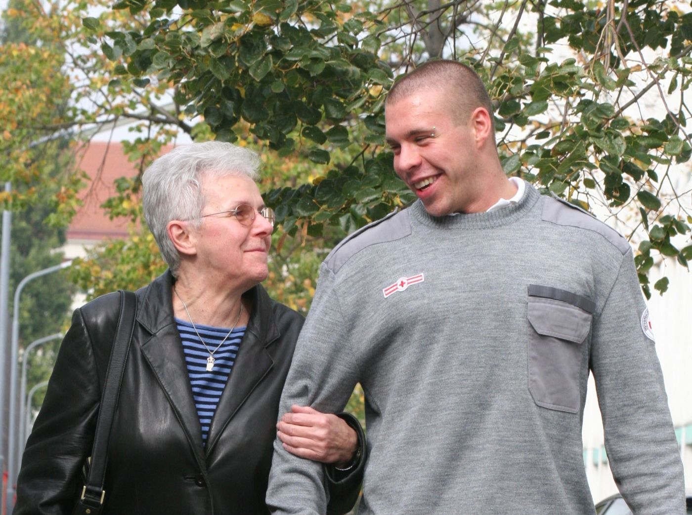 Ein Zivildiener in einer Uniform des Österreichischen Roten Kreuzes begleitet eine ältere Dame bei einem Spaziergang. Der Zivildiener trägt dabei eine Tasche.