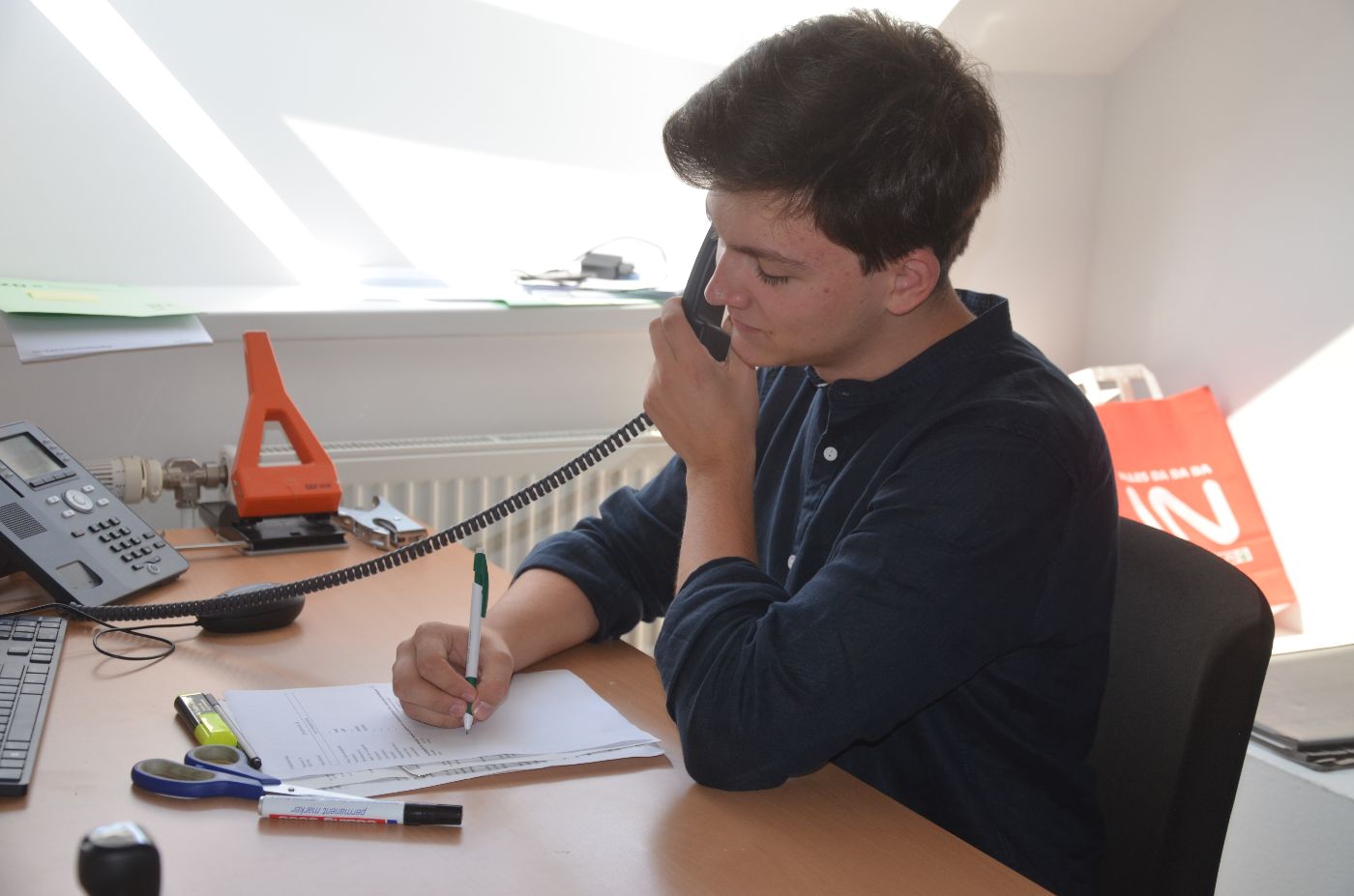 Ein Zivildiener sitzt an einem Schreibtisch, mit einer Hand hält er einen Telefonhörer an sein Ohr, mit der anderen Hand macht er Notizen
