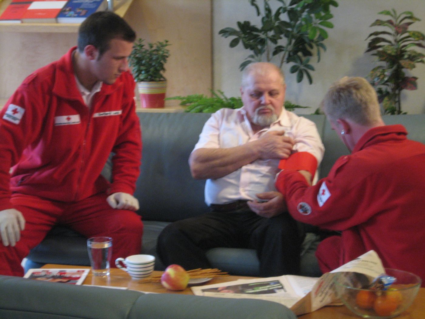 Zwei Zivildiener und älterer Mann, einer der beiden Zivildiener misst den Blutdruck des älteren Mannes