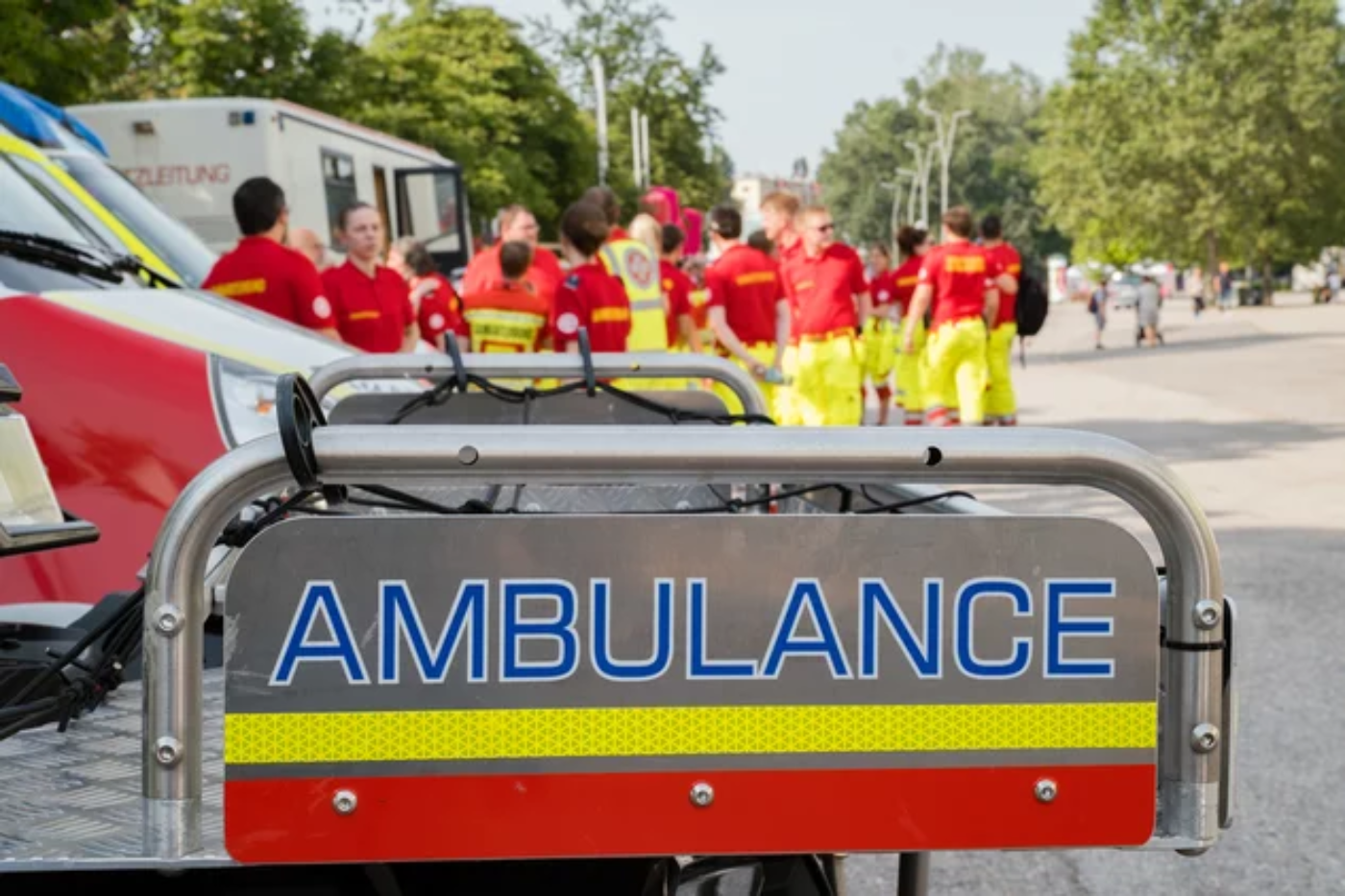 Ein Schild mit Aufdruck "Ambulance" in Großbuchstaben, im Hintergrund sind Zivildiener in Uniform des Samariterbundes Österreichs zu sehen