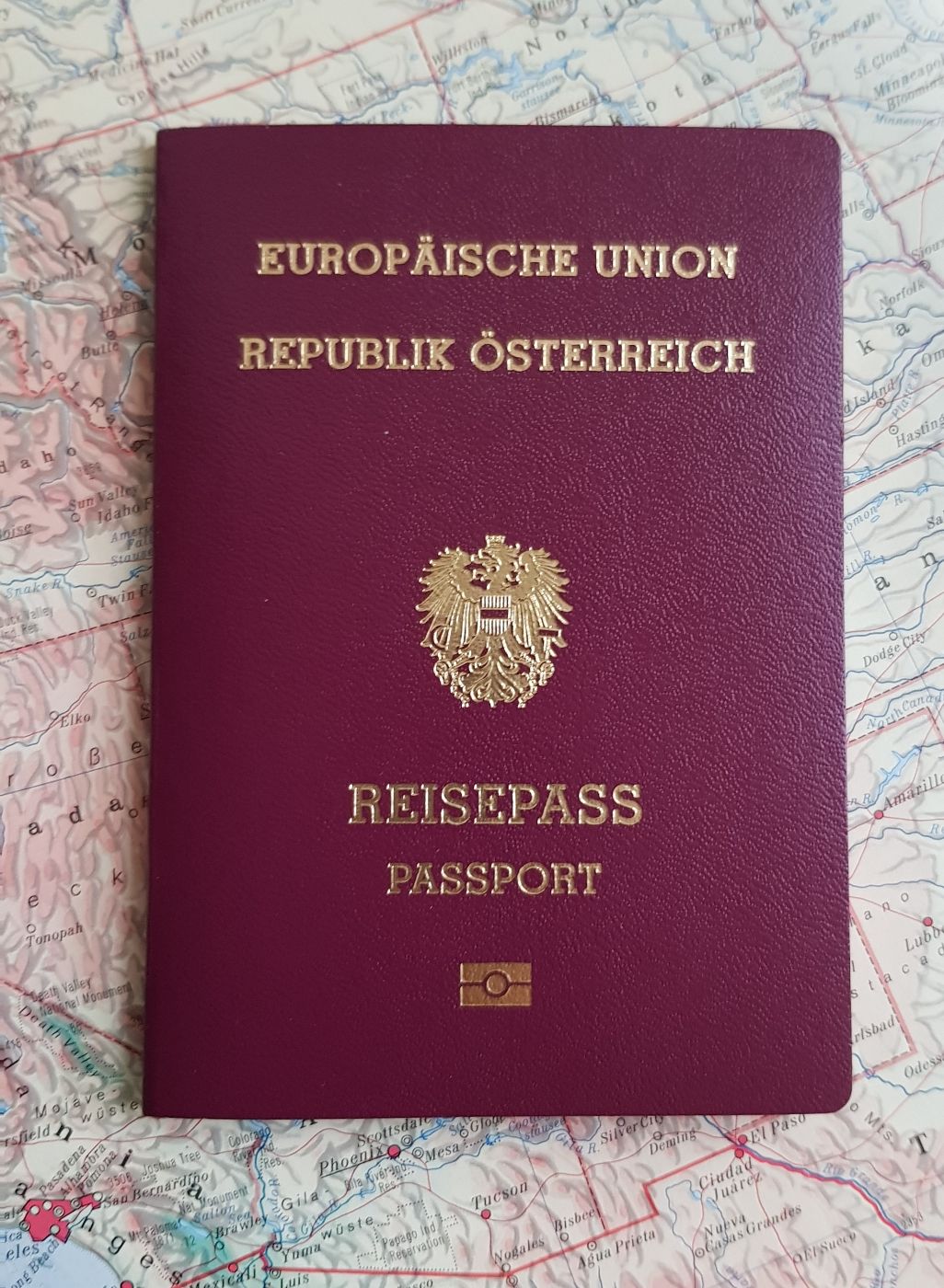 Abbildung des österreichischen Reisepasses. Der Reisepass liegt auf einer Landkarte. Der österreichische Reisepass ist bordeauxrot, in der Mitte ist das österreichische Bundeswappen in gold aufgedruckt, die Schrift ist ebenfalls in gold. Der Reisepass ist circa 100 mm breit und circa 160 mm hoch.