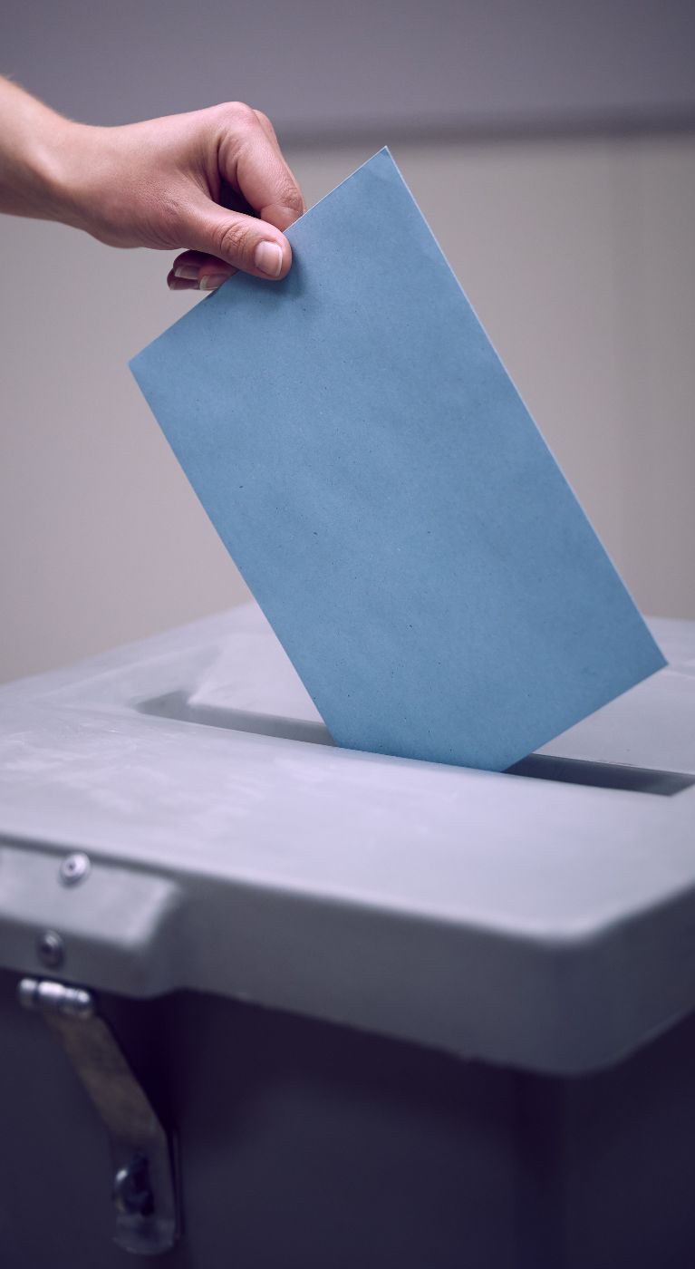 Das Bild zeigt die Hand einer Person, die ein blaues Kuvert in eine Wahlurne einwirft.