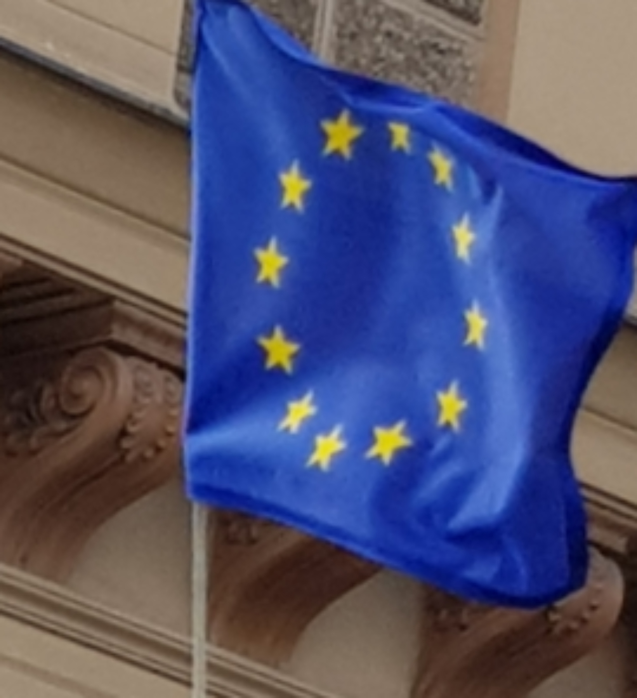 Flagge der Europäischen Union, Blau mit gelben Sternen; im Hintergrund ist ein Teil einer Gebäudefassade zu sehen
