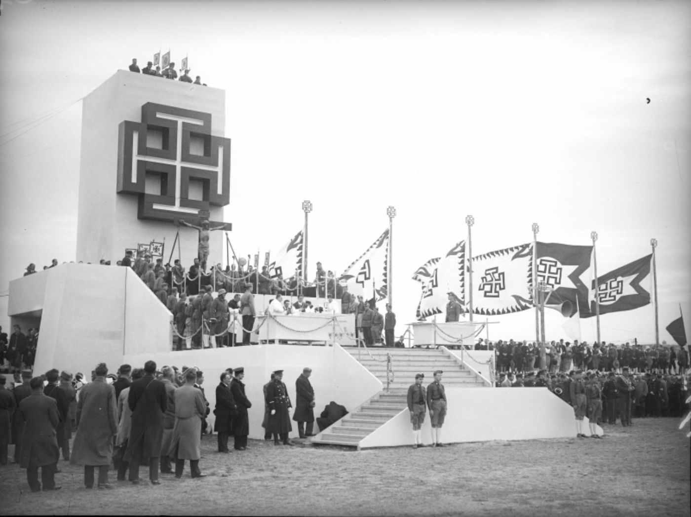 Abbildung eines Großappells der Vaterländischen Front zur Illustration. Das schwarz-weiß-Foto zeigt eine Rednertribüne mit Turm und Kruckenkreuz, mehrere Fahnen und viele Menschen.