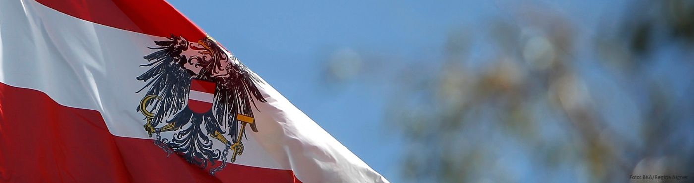 Bildnachweis: BKA/Regina Aigner; Österreichische Fahne in Rot-Weiß-Rot weht im Wind, im Hintergrund sind Äste eines Baumes und der Himmel zu sehen