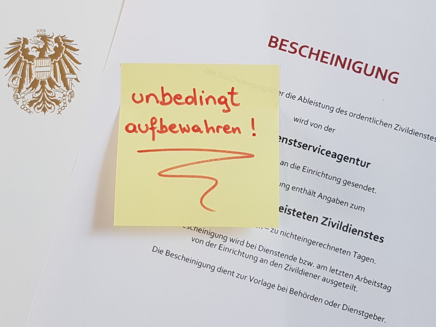 Ausschnitt eines Muster-Dokuments mit dem Titel „Bescheinigung“. Auf diesem klebt ein gelbes Post-It mit dem handgeschriebenen Aufdruck „unbedingt aufbewahren!“. Links im Bild befindet sich ein Ausschnitt einer Mappe, auf der der österreichische Bundesadler in der Farbe Gold aufgedruckt ist.
