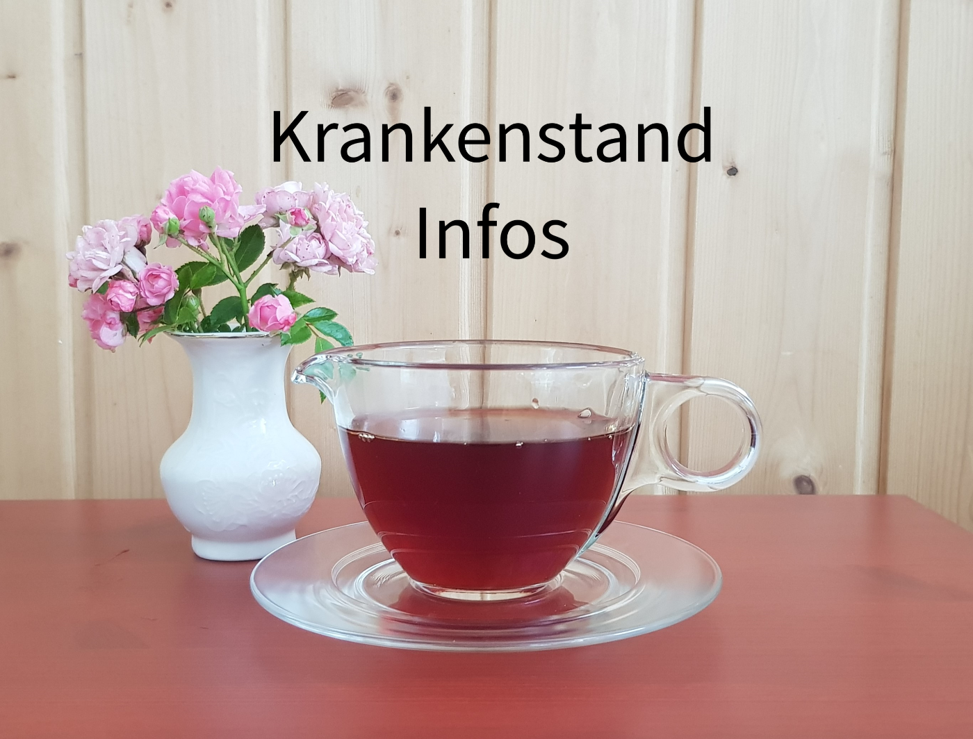 Symbolfoto mit einer Tasse aus Glas mit Tee, links daneben steht eine weiße Vase mit rosaroten Blumen. Die Tasse Tee steht auf einem roten Tisch.