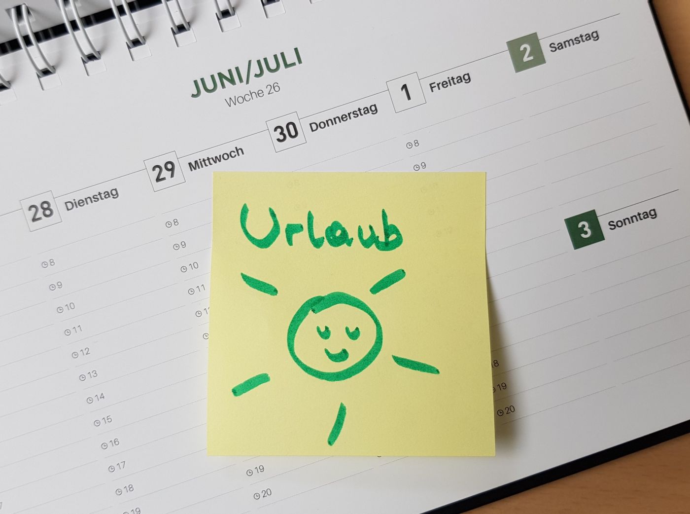 Kalenderblatt mit Überschrift Juni/Juli, auf dem Kalenderblatt klebt ein gelbes Post-It mit der Aufschrift "Urlaub" und einer aufgemalten lachenden Sonne