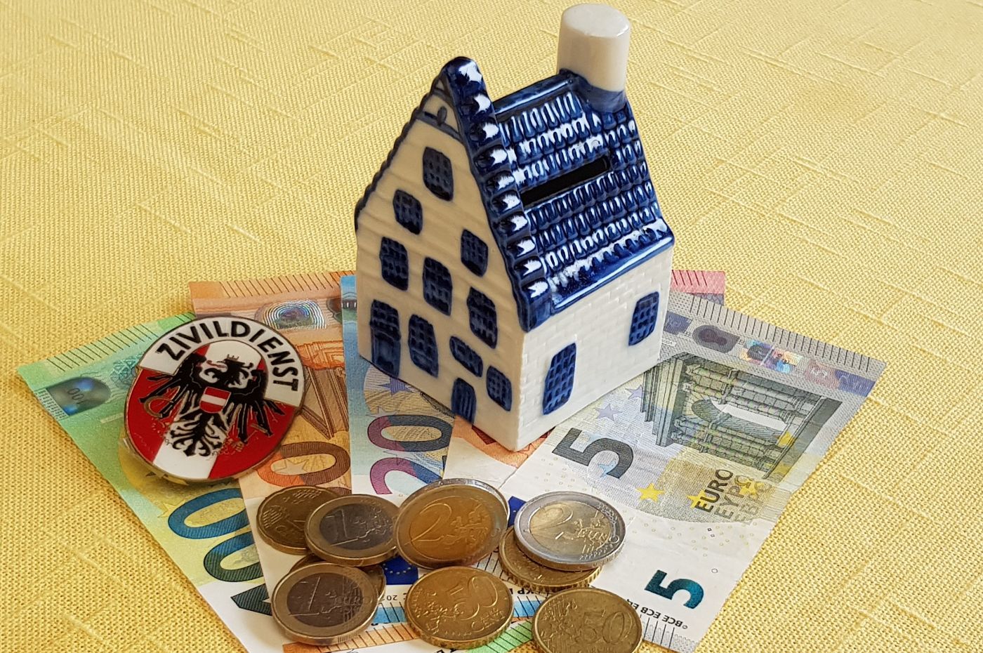 Kleines Keramik-Spielzeughaus steht auf Geldscheinen. Auf den Geldscheinen liegen Münzen. Die Fassade und der Rauchfang des Hauses sind weiß, die Tür, Fenster und das Dach sind blau.