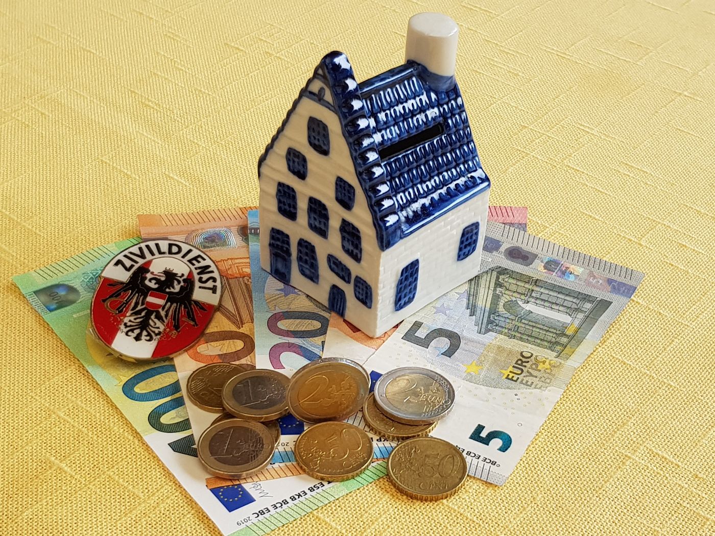 Kleines Keramik-Spielzeughaus steht auf Geldscheinen. Auf den Geldscheinen liegen Münzen. Die Fassade und der Rauchfang des Hauses sind weiß, die Tür, Fenster und das Dach sind blau.